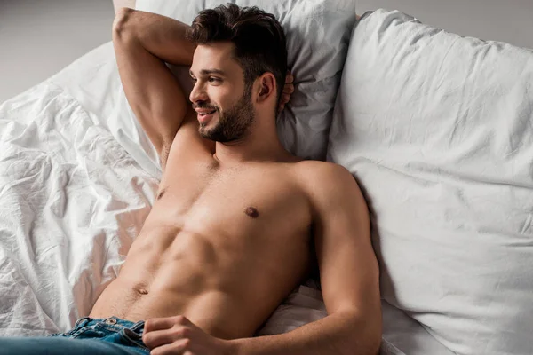 Улыбающийся сексуальный мачо без рубашки в джинсах лежащий на кровати на сером — стоковое фото