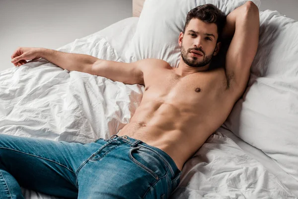 Schöner sexy hemdloser Mann in Jeans auf dem Bett liegend auf grau — Stockfoto