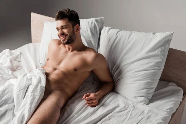 Sexy nu sorrindo homem deitado na cama no cinza — Fotografia de Stock
