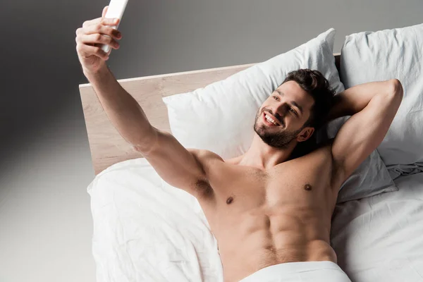 Alegre sin camisa sexy hombre tomando selfie en smartphone en la cama en gris - foto de stock