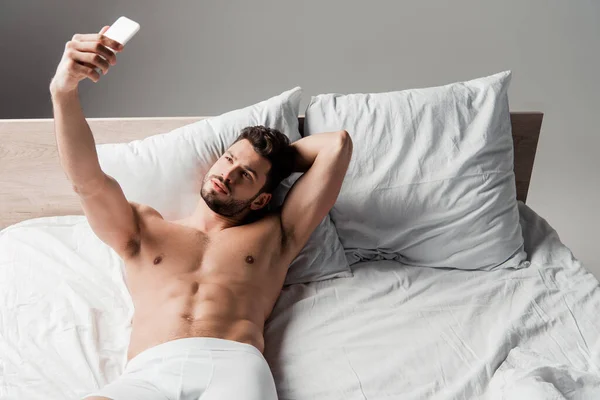 Красивий без сорочки сексуальний чоловік бере селфі на смартфон у ліжку на сірому — стокове фото