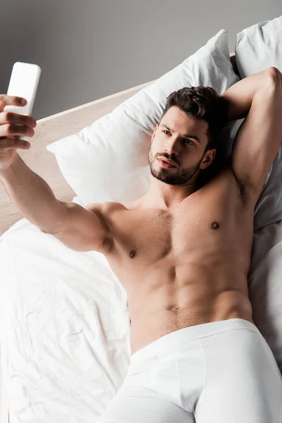 Shirtless sexy macho taking selfie on smartphone in bed on grey — Stock Photo