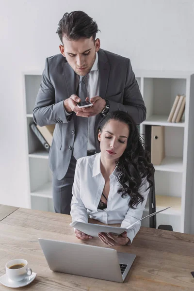 Handsome businessman taking photo of secretary sitting at workplace in unbuttoned blouse — Stock Photo