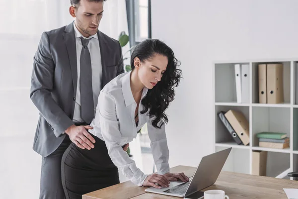 Guapo empresario tocando las caderas de la secretaria que trabaja en el portátil - foto de stock