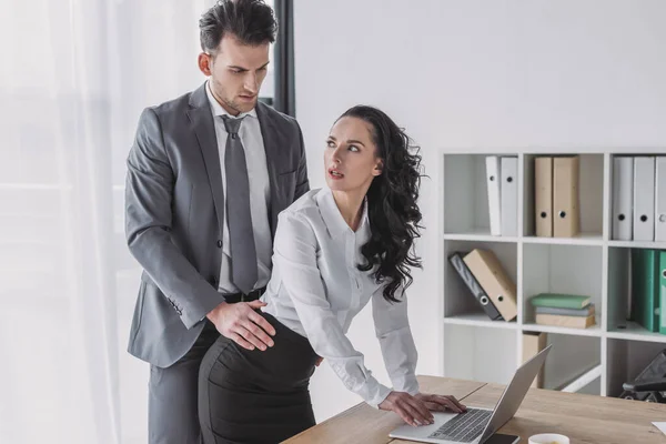 Handsome businessman touching hip of surprised secretary standing at workplace — Stock Photo