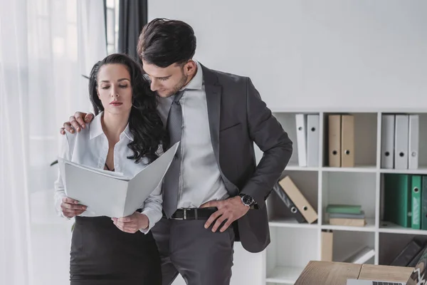 Guapo hombre de negocios tocando hombro de hermosa secretaria sosteniendo papeles - foto de stock