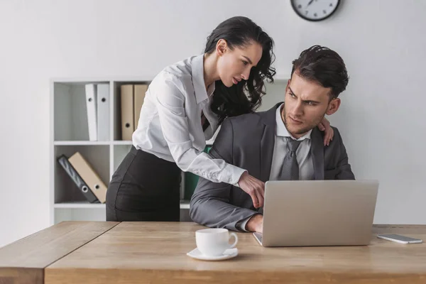 Secretaria sexy tocando hombro de empresario serio mientras que señala a la computadora portátil - foto de stock