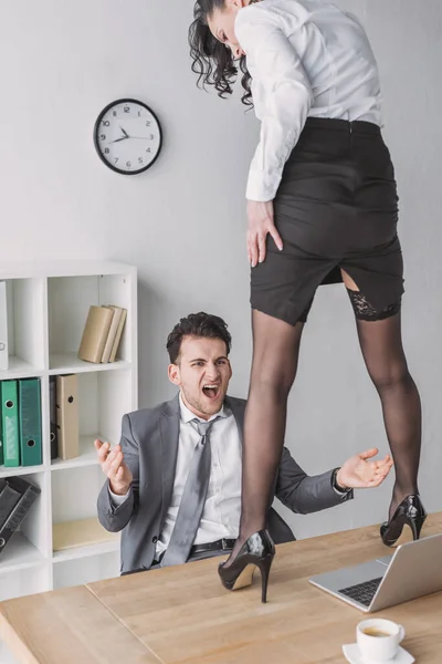 Hombre de negocios enojado gritando mientras mira a secretaria sexy de pie en el escritorio en zapatos de tacón alto - foto de stock