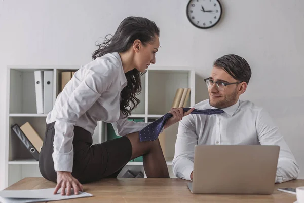 Sexy Geschäftsfrau sitzt am Schreibtisch und rührt Krawatte von lächelnden Kollegen, während sie ihn im Büro verführt — Stockfoto