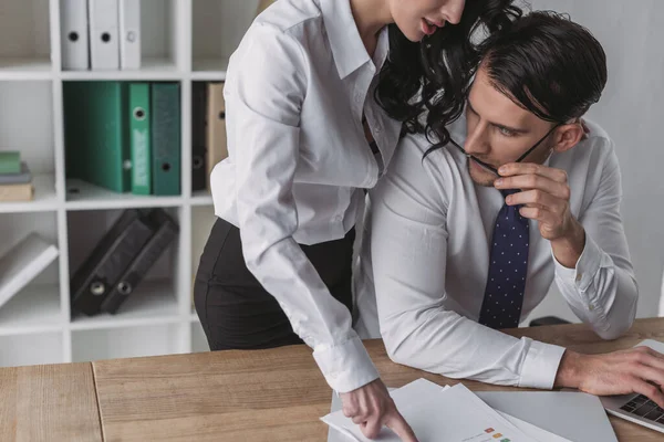 Sexy businesswoman pointing with finger at papers while standing close to colleague sitting at workplace — Stock Photo