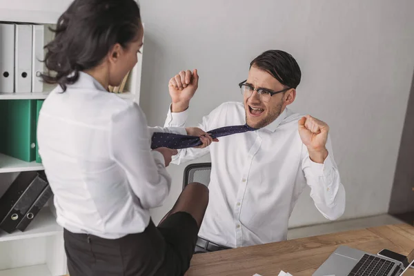 Sexy businesswoman sitting on desk and touching tie of shocked colleague while seducing him in office — Stock Photo