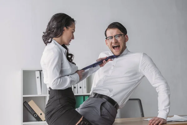 Passionate businesswoman touching tie of shocked colleague while seductive him in office — Stock Photo