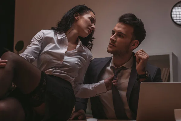 Selective focus of sexy secretary sitting on desk and touching tie of handsome businessman — Stock Photo