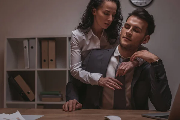 Atractiva, sensual secretaria abrazando hombre de negocios sentado en el escritorio con los ojos cerrados - foto de stock