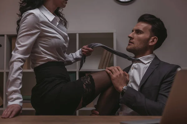 Cropped view of seductive secretary sitting on desk and touching tie of handsome businessman — Stock Photo