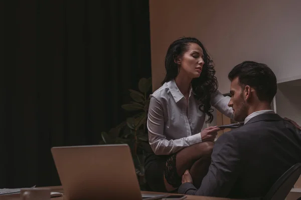 Seductive secretary sitting on desk and touching tie of businessman in formal wear — Stock Photo