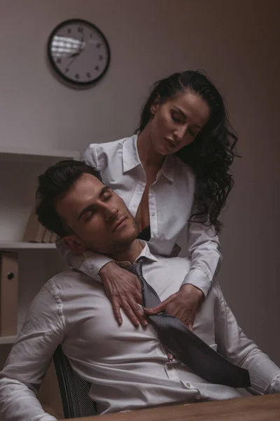 Apasionada secretaria en blusa desabotonada abrazando guapo hombre de negocios sentado con los ojos cerrados - foto de stock