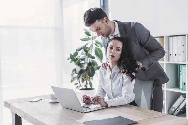 Handsome businessman touching shoulders of disappointed secretary sitting at workplace — Stock Photo