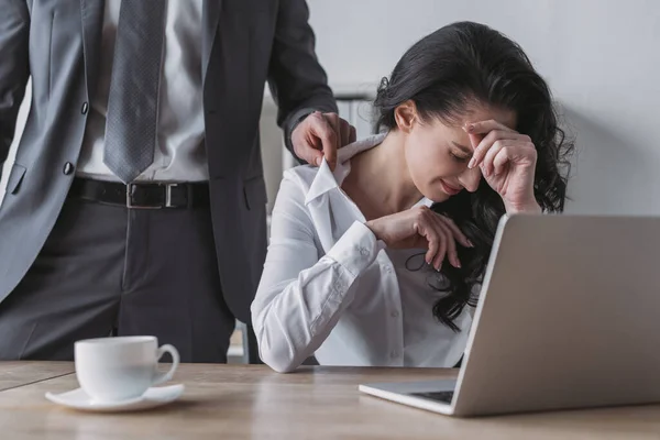 Abgeschnittene Ansicht des Geschäftsmannes, der die Bluse einer unzufriedenen Sekretärin berührt, die am Arbeitsplatz sitzt und den Kopf berührt — Stockfoto