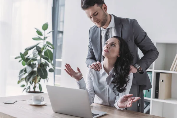 Handsome businessman touching shoulders of displeased secretary sitting at workplace — Stock Photo