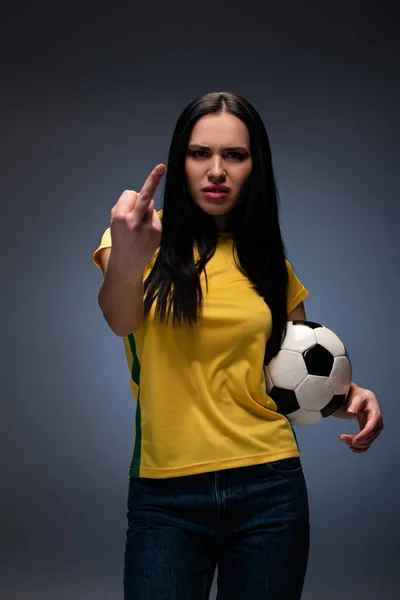 Agresivo ventilador de fútbol femenino sosteniendo la pelota mientras muestra el dedo medio en gris - foto de stock