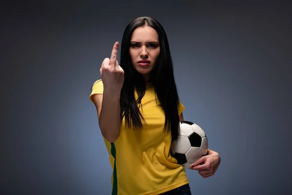 Fan de football féminin en colère tenant le ballon tout en montrant le majeur sur gris — Photo de stock
