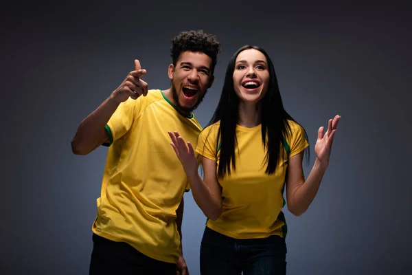Emocional pareja multiétnica de aficionados al fútbol en camisetas amarillas gestos en gris - foto de stock