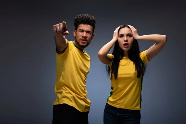 Preocupados pareja multiétnica de aficionados al fútbol en camisetas amarillas apuntando en gris - foto de stock