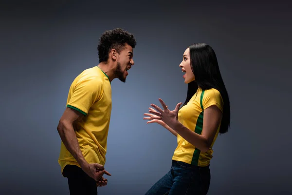 Multiethnic couple of emotional excited football fans in yellow t-shirts shouting on grey — Stock Photo