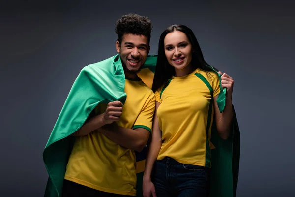 Pareja multiétnica de aficionados al fútbol feliz con bandera de Brasil en gris - foto de stock