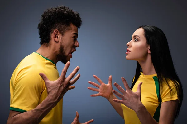 Agressivo casal multicultural de fãs de futebol em camisetas amarelas gesticulando e gritando em cinza — Fotografia de Stock