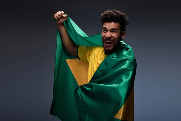 Усміхнений афроамериканський футбольний фанат, загорнутий у бразильський прапор на сірому — стокове фото