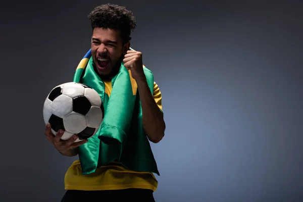 Fröhlicher afrikanisch-amerikanischer Mann mit brasilianischer Flagge brüllt und hält Fußballball auf grau — Stockfoto