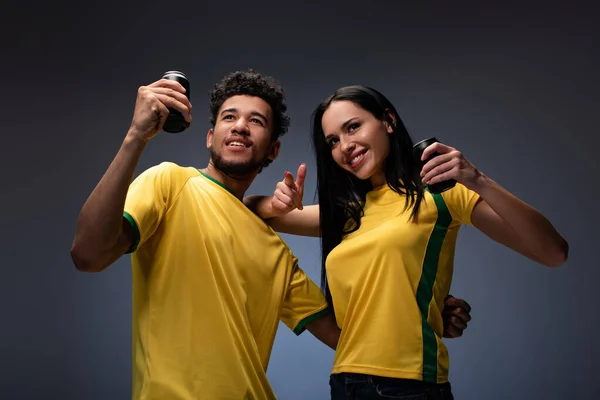 Multiethnic couple of smiling football fans in yellow t-shirts holding cans with drink on grey — Stock Photo