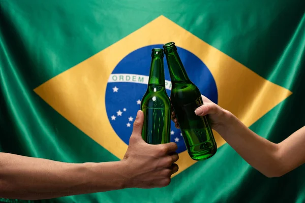 Cropped view of couple of football fans clinking with bottles of beer with brazil flag behind — Stock Photo