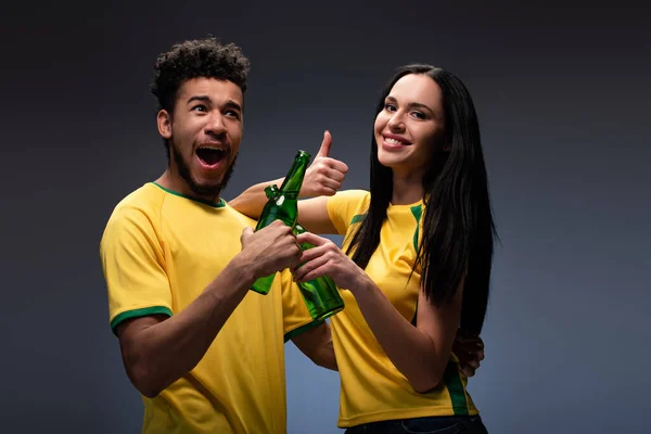 Excited multiethnic couple of football fans in yellow t-shirts clinking with bottles of beer and showing thumb up on grey — Stock Photo