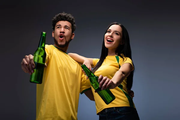 Excitada pareja multiétnica de aficionados al fútbol en camisetas amarillas sosteniendo botellas de cerveza en gris - foto de stock