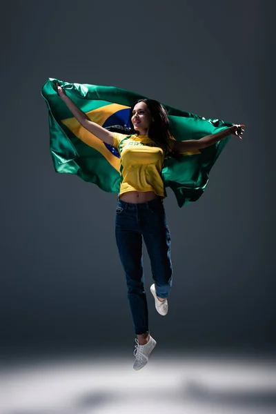 Attractive cheerful football fan jumping with brazilian flag on grey — Stock Photo