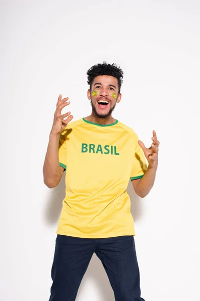 Emocionado fanático del fútbol afroamericano en camiseta amarilla con signo brasileño gestos y de pie sobre gris - foto de stock
