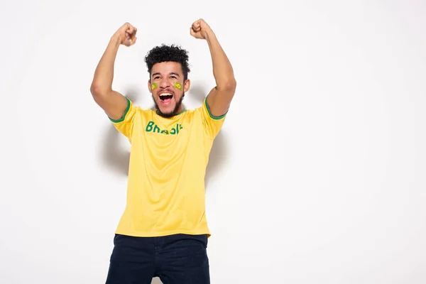 Emocionado fanático del fútbol afroamericano en camiseta amarilla con signo brasileño gestos y gritos en gris - foto de stock