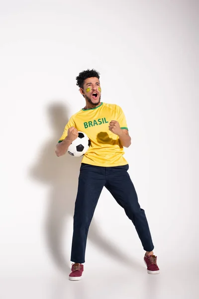 Емоційний афроамериканський футбольний фанат у жовтій футболці кричить і тримає м'яч на сірому — стокове фото