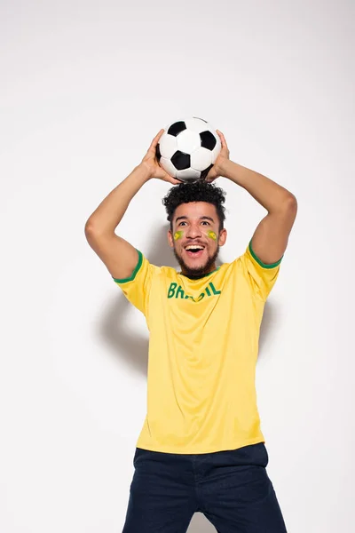 Emocionado fanático del fútbol afroamericano en camiseta amarilla sosteniendo la pelota en gris - foto de stock
