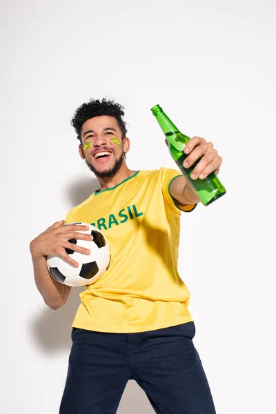 Uomo afroamericano sorridente che tiene palla da calcio e bottiglia di birra sul grigio — Foto stock