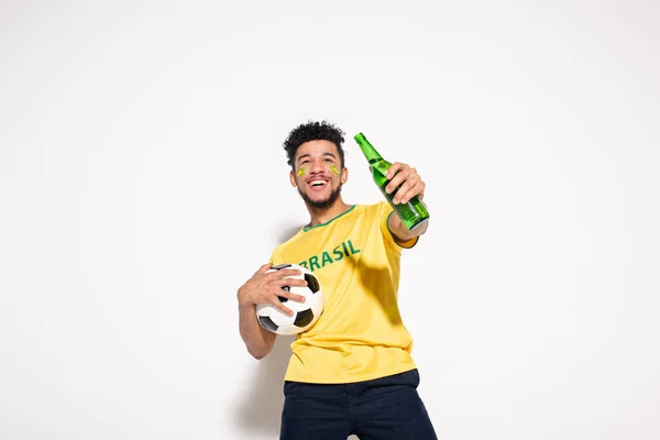 Homem americano africano alegre segurando bola de futebol e garrafa de cerveja em cinza — Fotografia de Stock