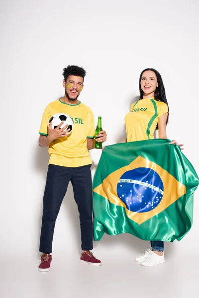 Multicultural couple of happy football fans holding brazilian flag, ball and bottle of beer on grey — Stock Photo