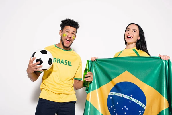 Multiethnic couple of excited football fans holding brazilian flag, ball and bottle of beer on grey — Stock Photo