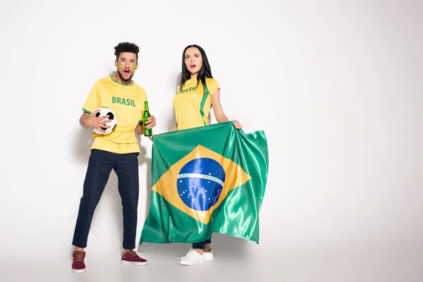 Multiethnic couple of surprised football fans holding brazilian flag, ball and bottle of beer on grey — Stock Photo