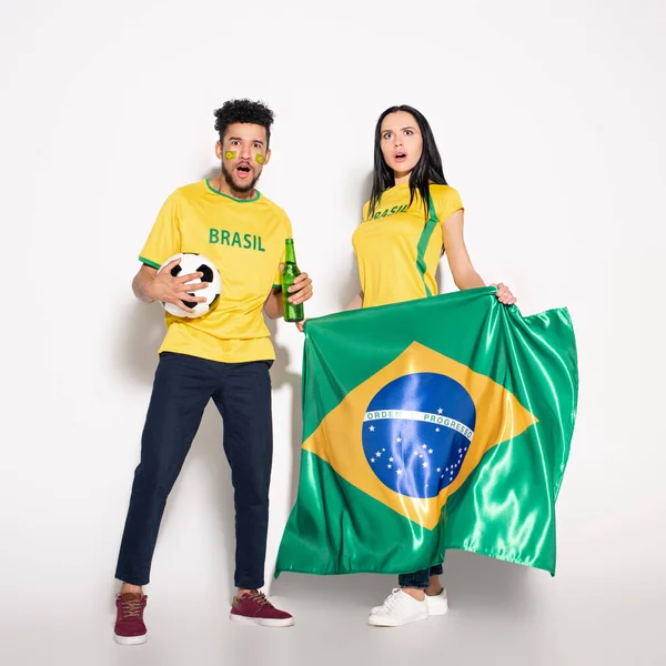 Multiethnic couple of shocked football fans holding brazilian flag, ball and bottle of beer on grey — Stock Photo