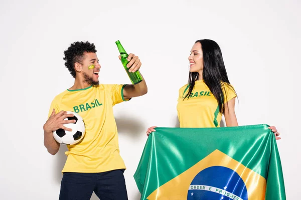 Casal multicultural de fãs de futebol feliz segurando bandeira brasileira, bola e garrafa de cerveja em cinza — Fotografia de Stock