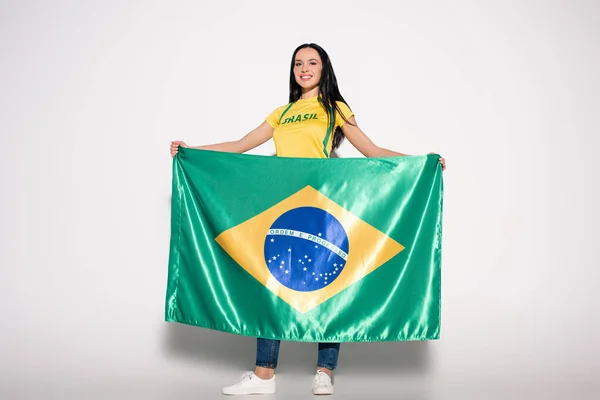 Atractiva y sonriente fan del fútbol femenino sosteniendo bandera brasileña en gris - foto de stock